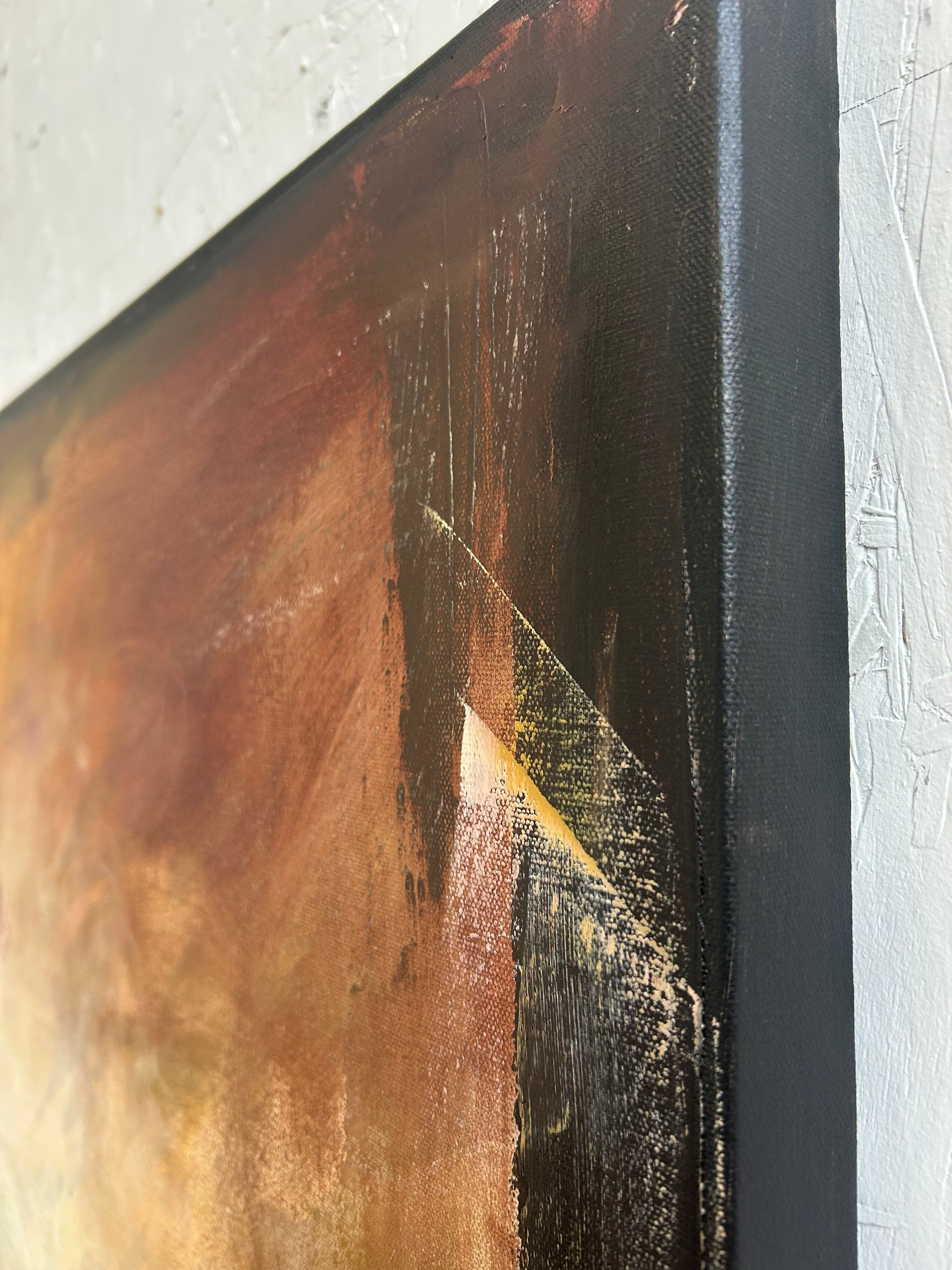 Close-up of a textured painting with gold and black elements on a white wall.