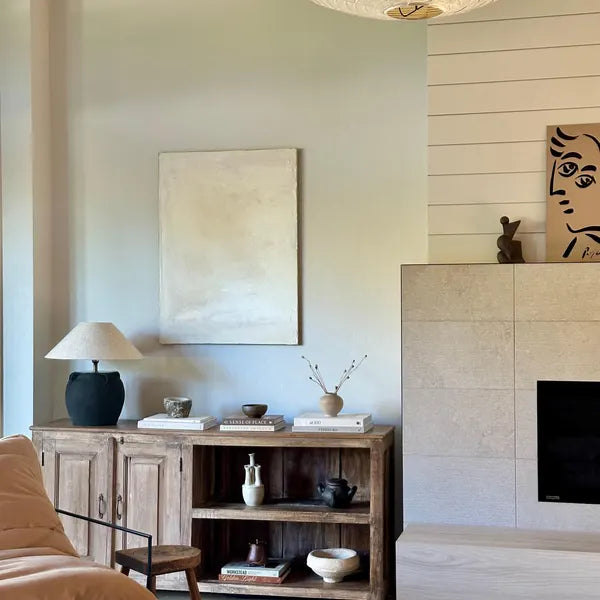 A minimalist white abstract painting displayed on a wall above a wooden console table in a room with a fireplace and a lamp.