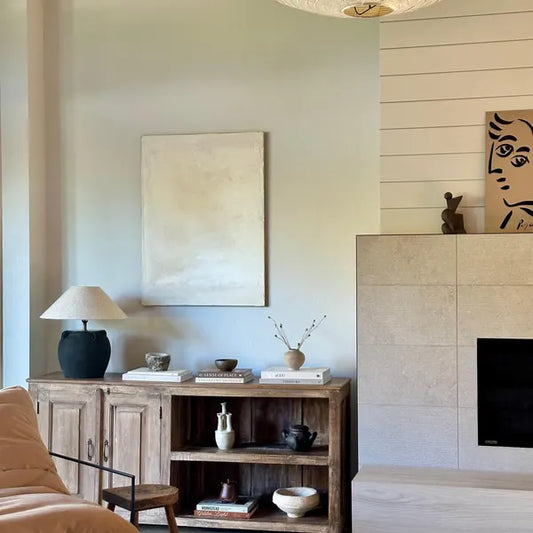 A minimalist white abstract painting displayed on a wall above a wooden console table in a room with a fireplace and a lamp.